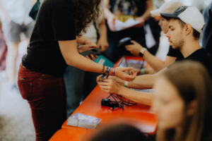 Eine Person übergibt ein Armband an einen Mitarbeiter am Empfangstisch des Elbenwald Festivals, umgeben von weiteren Besuchern und Mitarbeitern. Der Tisch ist mit Programmheften und technischen Geräten ausgestattet.