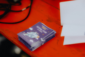 Stapel von Festival-Atlas-Heften auf einem orangefarbenen Tisch, die Informationen zum Elbenwald Festival enthalten. Die Hefte sind lila mit bunten Illustrationen, die die nerdige Atmosphäre des Festivals widerspiegeln.