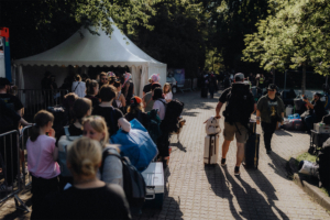 Menschenmenge vor einem Zelt beim Elbenwald Festival, das eine Mischung aus Musikfestival und Convention bietet. Teilnehmer in bunten Outfits und mit Gepäck warten in der Schlange, um die Veranstaltung zu betreten. Sonniger Tag mit viel Grün im Hintergrund.