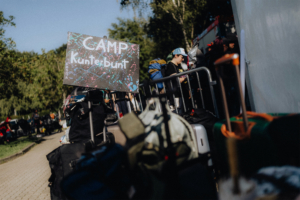 Campingbereich "Kunterbunt" beim Elbenwald Festival, umgeben von Gepäck und Teilnehmern. Bunte Schilder und eine lebhafte Atmosphäre prägen das Festivalgelände, das Musik und Nerd-Kultur vereint.