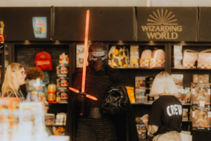 Ein verkleideter Besucher mit rotem Lichtschwert und Maske steht an einem Verkaufsstand auf dem Elbenwald Festival. Im Hintergrund sind Merchandise-Artikel und bunte Rucksäcke zu sehen, während zwei Frauen mit einem Crew-Shirt und einer Brille miteinander sprechen.