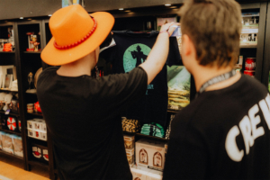 Zwei Besucher des Elbenwald Festivals betrachten ein T-Shirt mit einem grafischen Design. Im Hintergrund sind verschiedene Merchandise-Artikel und Poster aus der Nerd-Kultur zu sehen, die das kreative Ambiente des Festivals unterstreichen.