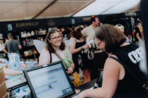 Besucherinnen des Elbenwald Festivals stehen am Merchandise-Stand. Eine Frau mit Feenflügeln lächelt, während sie mit einer Verkäuferin spricht. Im Hintergrund sind zahlreiche Festivalbesucher und bunte Merchandise-Artikel zu sehen.