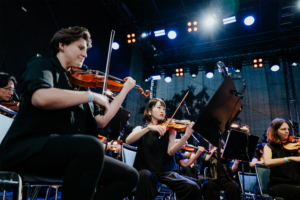 Elbenwald in Concert Ein Orchester spielt live auf der Bühne des Elbenwald Festivals, während die Musikerinnen und Musiker in schwarzer Kleidung konzentriert ihre Violinen spielen. Bunte Lichter und eine lebendige Atmosphäre schaffen eine einzigartige Festivalstimmung.