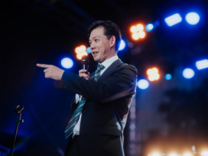 Elbenwald in Concert Ein Redner in einem Anzug hält ein Mikrofon und spricht enthusiastisch auf der Bühne des Elbenwald Festivals, umgeben von bunten Lichtern und einer lebhaften Atmosphäre, die Musik und Nerdkultur vereint.