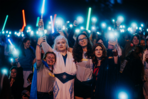Elbenwald in Concert: Eine Gruppe von vier Frauen posiert lächelnd auf dem Elbenwald Festival, während sie leuchtende Lichtschwerter in der Hand halten. Die Menge im Hintergrund ist mit bunten Lichtern erleuchtet, was die festliche und lebendige Atmosphäre des Events unterstreicht. Foto: Robin Schmiedebach