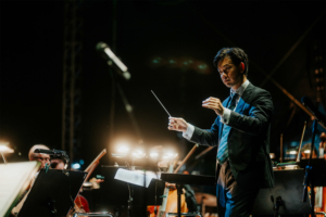 Elbenwald in Concert Dirigent leitet ein Orchester während des Elbenwald Festivals, umgeben von Musiker*innen und Bühnenlicht. Die Atmosphäre verbindet Musik und Nerd-Kultur in einem einzigartigen Erlebnis.
