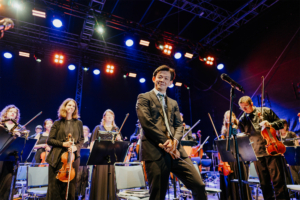 Elbenwald in Concert: Ein Musiker in Anzug steht im Vordergrund einer Orchestervorstellung beim Elbenwald Festival. Im Hintergrund sind weitere Orchestermitglieder mit Geigen und Notenständern zu sehen, umgeben von bunten Lichtern auf der Bühne. Foto: Robin Schmiedebach