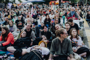 Eine große Menschenmenge sitzt entspannt auf dem Boden beim Elbenwald Festival, umgeben von bunten Lichtern und fröhlichen Gesichtern. Die Teilnehmer genießen die Atmosphäre und die Musik, während sie sich in ihren nerdigen Outfits präsentieren.
