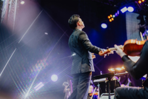 Elbenwald in Concert Ein Dirigent in eleganter Kleidung leitet ein Orchester auf der Bühne des Elbenwald Festivals, umgeben von bunten Lichtern und einer energiegeladenen Atmosphäre, die Musik und Nerdkultur vereint.