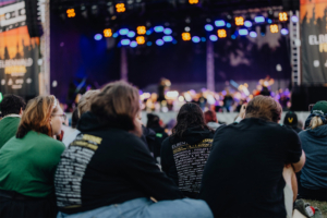 Teilnehmer des Elbenwald Festivals genießen eine musikalische Darbietung, während sie auf dem Gelände sitzen. Im Hintergrund sind die bunten Lichter der Bühne und das Festival-Logo sichtbar, was die festliche Atmosphäre unterstreicht.