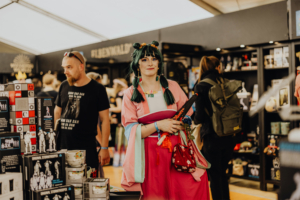 Frau im bunten Kostüm mit grünen Haaren hält eine Spielzeugwaffe und Blumen in einem Verkaufsstand auf dem Elbenwald Festival, umgeben von Merchandise-Artikeln und anderen Besuchern. Die Atmosphäre verbindet Musik, Kunst und Nerd-Kultur.