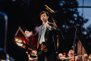 Elbenwald in Concert Dirigent bei Elbenwald Festival, umgeben von Orchestermitgliedern. Die Atmosphäre ist lebhaft, während er mit Begeisterung das Konzert leitet. Die Bühne ist beleuchtet und die Musiker sind bereit, ihre Darbietung zu beginnen.