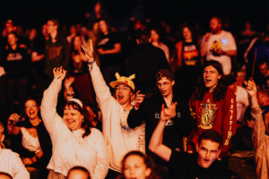 Menschenmenge beim Elbenwald Festival, die begeistert feiert. Teilnehmer in bunten Kostümen und T-Shirts zeigen ihre Freude, während sie zur Musik tanzen. Die Atmosphäre ist lebhaft und voller Energie, mit einer Mischung aus Fans aus verschiedenen Nerd-Kulturen.