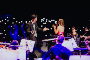 Elbenwald in Concert Ein Dirigent leitet ein Orchester während des Elbenwald Festivals, während eine Sängerin im roten Kleid das Publikum mit ihrem Gesang begeistert. Im Hintergrund leuchten zahlreiche Handys, die die Atmosphäre des Festivals unterstreichen.