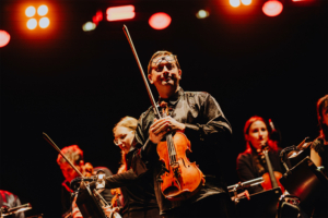 Elbenwald in Concert Ein Musiker hält eine Geige auf der Bühne des Elbenwald Festivals, umgeben von anderen Orchestermusikern. Die Lichter strahlen in warmen Rottönen und schaffen eine lebendige Atmosphäre für das Publikum.