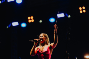 Elbenwald in Concert Sängerin auf der Bühne des Elbenwald Festivals, umgeben von bunten Lichtern, während sie ein Mikrofon hält und mit einem Taktstock dirigiert. Die Atmosphäre ist lebhaft und energiegeladen, perfekt für Fans von Musik und Nerdkultur.