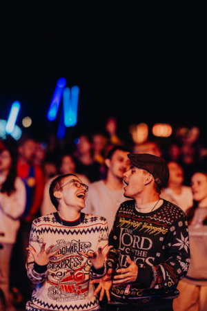 Zwei fröhliche Festivalbesucher in nerdigen Pullovern stehen inmitten einer jubelnden Menge beim Elbenwald Festival, umgeben von bunten Lichtern. Die Atmosphäre ist lebhaft und voller Begeisterung für Musik und Fantasie.
