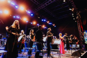Elbenwald in Concert Auf der Bühne des Elbenwald Festivals steht ein Orchester mit Musikern in eleganter Kleidung, die Instrumente halten. Bunte Lichter strahlen auf die Darbietung, während das Publikum applaudiert und die Atmosphäre festlich und begeistert ist.