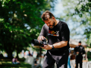 eosAndy Ein Fotograf in einem schwarzen T-Shirt mit dem Aufdruck "Gen-X" konzentriert sich auf seine Kamera während des Elbenwald Festivals. Umgeben von grüner Natur und Festivalbesuchern, fängt er die lebendige Atmosphäre des Events ein.