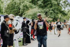 Ein fröhlicher Festivalbesucher posiert vor einer Gruppe von Cosplayern in kreativen Kostümen beim Elbenwald Festival, umgeben von Bäumen und Festivalatmosphäre. Die Stimmung ist lebhaft und bunt, ideal für Fans von Popkultur und Musik.