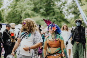 Bunt gekleidete Festivalbesucher beim Elbenwald Festival, das Musik und Cosplay vereint. Im Vordergrund sind ein Mann mit lockigem Haar und eine Frau mit bunten Haaren und Blumen im Haar zu sehen, während im Hintergrund weitere verkleidete Gäste erscheinen.