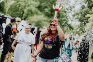 Eine fröhliche Teilnehmerin des Elbenwald Festivals hält eine bunte Blume in der Hand, während Seifenblasen um sie herum schweben. Im Hintergrund sind weitere Festivalbesucher in kreativen Kostümen zu sehen, die die lebendige Atmosphäre des Events unterstreichen.