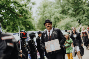 Ein Mann im Anzug und Zylinder hält ein Schild, während er auf dem Elbenwald Festival mit einer Gruppe von Cosplayern, darunter eine Figur im Link-Kostüm, an einem grünen Ort läuft. Die Atmosphäre ist lebhaft und bunt, typisch für ein nerdiges Event.