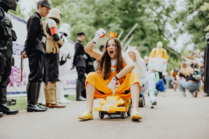 Ein fröhliches Kind in einem orangefarbenen Kostüm sitzt auf einem gelben Spielzeugauto und posiert enthusiastisch, umgeben von Cosplayern in verschiedenen Kostümen beim Elbenwald Festival, das eine Mischung aus Musikfestival und Convention bietet.