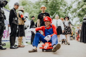Ein fröhlicher Teilnehmer im Mario-Kostüm fährt in einem kleinen roten Kart beim Elbenwald Festival. Im Hintergrund sind verschiedene Cosplayer in beeindruckenden Kostümen zu sehen, die die bunte Atmosphäre des Events unterstreichen.