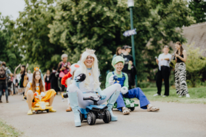 Kostümierte Teilnehmer des Elbenwald Festivals fahren in Mini-Autos auf einem Weg. Zu sehen sind Figuren in bunten Outfits, darunter Prinzessin Peach und Luigi, umgeben von einer fröhlichen Festivalatmosphäre und Zuschauern im Hintergrund.