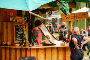 Bunt dekorierter Waffelstand auf dem Elbenwald Festival, mit einem Drachenkopf und einem Menü, das verschiedene Waffelvariationen anbietet. Besucher genießen die entspannte Atmosphäre und das kreative Ambiente des Festivals.
