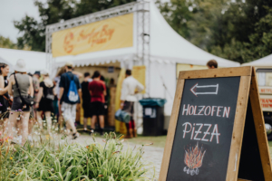 Ein Holzofen-Pizza-Schild zeigt den Weg zu einem Essensstand beim Elbenwald Festival. Im Hintergrund sind Besucher in festlicher Kleidung zu sehen, die die lebendige Atmosphäre des Festivals genießen. Blumen und Gräser schmücken den Vordergrund.