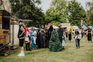 Besucher in fantasievollen Kostümen stehen an einem Food Truck beim Elbenwald Festival, umgeben von bunten Ständen und einer lebhaften Festivalatmosphäre. Eine festliche Dekoration in Form eines Weihnachtsbaums ist ebenfalls zu sehen.