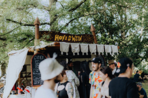 Food Bunte Menschenmenge vor einem Honigweinstand auf dem Elbenwald Festival, umgeben von Bäumen. Das Schild zeigt Getränkeangebote wie Met und Cider. Die festliche Atmosphäre und die kreative Dekoration schaffen eine einladende Umgebung für Festivalbesucher.