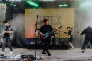 Gate X Live Musiker der Band Gate X treten live auf der Bühne beim Elbenwald Festival. Die Atmosphäre ist energiegeladen, mit bunten Lichtern und Nebel, während die Bandmitglieder intensiv spielen. Im Hintergrund sind Instrumente und Technik zu sehen.