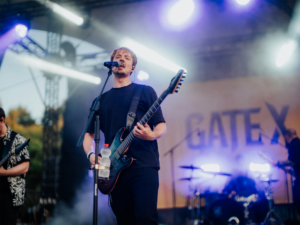 Gate X live Ein Musiker steht auf der Bühne des Elbenwald Festivals und spielt Gitarre, während das Publikum begeistert zuschaut. Im Hintergrund sind Bandmitglieder und Lichteffekte zu sehen, die die energiegeladene Atmosphäre des Events unterstreichen.