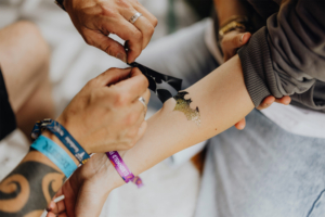 Glitzer-Tattoos Hände, die ein glitzerndes Bat-Symbol auf den Arm einer Person auftragen, umgeben von Festivalarmbändern. Die Szene fängt die kreative Atmosphäre des Elbenwald Festivals ein, das Musik und Nerdkultur vereint.