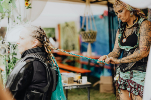 Haare flechten Eine Friseurin mit Tattoos und bunten Haaren flechtet Zöpfe für eine Teilnehmerin des Elbenwald Festivals, das Musik und Nerdkultur vereint. Im Hintergrund sind bunte Stände und Sitzgelegenheiten sichtbar, die zur festlichen Atmosphäre beitragen.