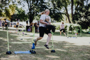 Hobby Horsing Teilnehmerin beim Elbenwald Festival springt mit einem Spielzeugpferd über Hindernisse. Die Veranstaltung kombiniert Musik und nerdige Aktivitäten im Freien, während andere Besucher im Hintergrund die Atmosphäre genießen.