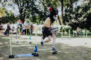 Hobby Horsing Ein Teilnehmer in einem Tierkostüm springt über Hindernisse beim Elbenwald Festival. Im Hintergrund sind weitere Festivalbesucher und bunte Attraktionen zu sehen. Die Atmosphäre ist lebhaft und kreativ, ideal für Fans von Popkultur und Musik.