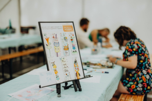 Creative Hobby Workshop Bunte Lesezeichen aus Aquarellfarben werden beim Elbenwald Festival gestaltet. Im Vordergrund steht ein Display mit kreativen Ideen, während Teilnehmer an Tischen ihre eigenen Designs kreieren. Preis pro Stück: 5 €.
