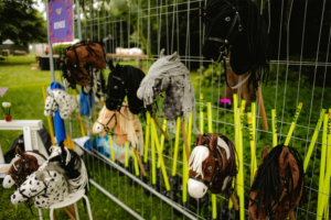 Hobby Horsing Bunte Pferdekostüme hängen an einem Zaun auf dem Elbenwald Festival, umgeben von leuchtenden Stäben. Die verschiedenen Modelle zeigen eine kreative Mischung aus verschiedenen Pferdearten, die die fantasievolle Atmosphäre des Festivals unterstreichen.