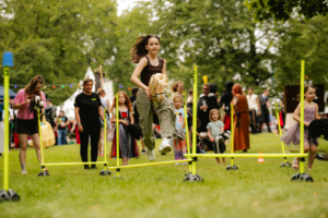 Hobby Horsing Ein Mädchen springt mit einem Spielzeugpferd über Hürden auf dem Elbenwald Festival. Im Hintergrund sind andere Kinder und Festivalbesucher in Kostümen zu sehen, die die lebhafte Atmosphäre des Events genießen.