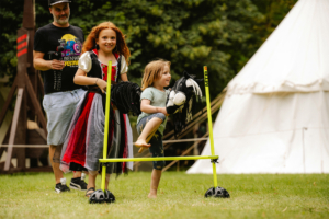 Hobby Horsing Zwei Kinder in Kostümen beim Elbenwald Festival, eines trägt ein langes, rotes Kleid und hält ein Spielzeugpferd, während das andere barfuß über einen Hindernisparcours springt. Im Hintergrund ist ein großes Zelt zu sehen.