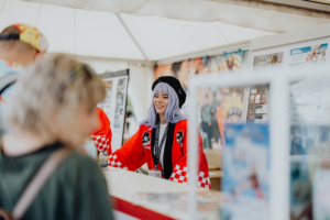 Ichiban Kuji Lotterie Lächelnde Cosplayerin mit lilafarbenem Haar und rotem Kimono während des Elbenwald Festivals, umgeben von Informationsständen. Die Atmosphäre vereint Musik und Nerd-Kultur, während Besucher die kreative Vielfalt des Events genießen.