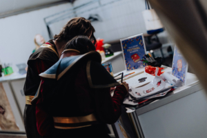 Infopoint Zwei Besucher des Elbenwald Festivals in kostümierten Outfits arbeiten an einem kreativen Projekt an einem Tisch, umgeben von Zeichenmaterialien und bunten Deko-Elementen. Im Hintergrund sind Festivalplakate sichtbar, die die Atmosphäre des Events unterstreichen.