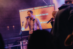 Ein Sänger auf der Bühne des Elbenwald Festivals, umgeben von einem begeisterten Publikum. Im Hintergrund leuchtet ein Bildschirm mit dem Text „Willkommen auf dem EWF25 - schön, dass“. Die Atmosphäre ist lebhaft und energiegeladen.