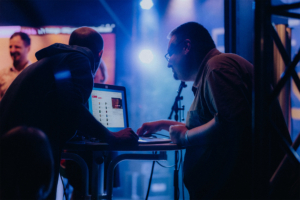 Zwei Personen arbeiten an einem Laptop während des Elbenwald Festivals. Die Szene ist von buntem Licht und einer lebhaften Atmosphäre geprägt, die die Mischung aus Musik und Nerdkultur widerspiegelt.