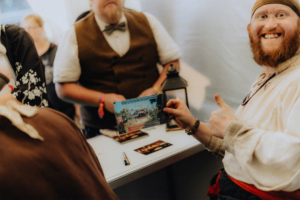 Meet and Greet Katerfahrt Ein Teilnehmer des Elbenwald Festivals hält begeistert eine Karte mit dem Titel „Käpt'n Kunolds Karpferfahrten“ in der Hand. Er trägt ein historisches Kostüm und zeigt Daumen hoch, während andere Besucher im Hintergrund zu sehen sind.